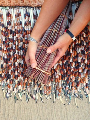 Hands holding reeds for basket weaving with shell necklaces.