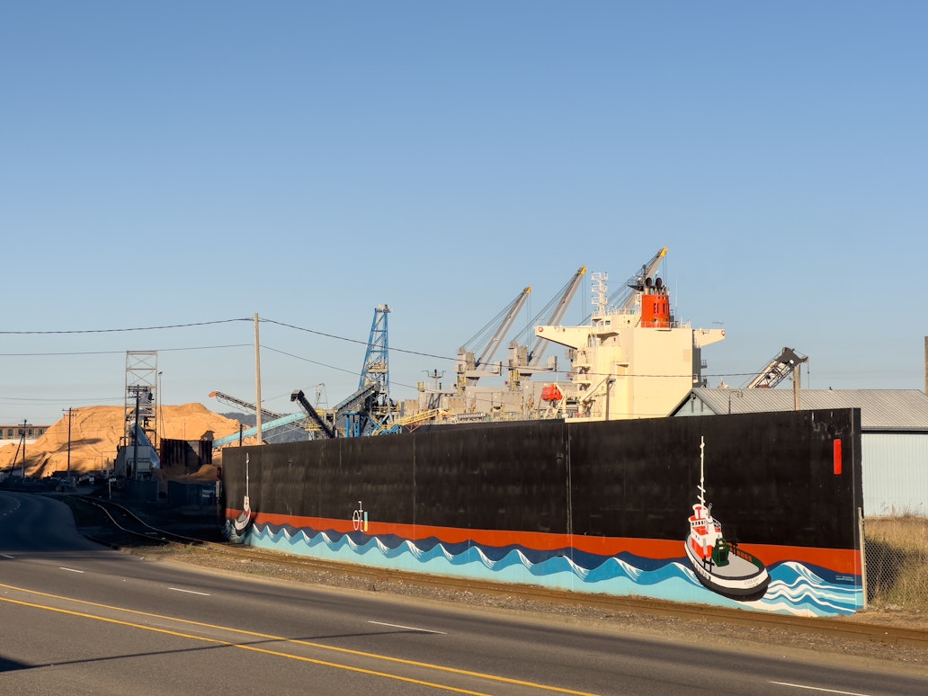 Wide view of the 101 Ship Wall Mural in the Empire District of Coos Bay showing painted tugboats riding blue waves along a long black wall, with an active industrial port and cargo ship visible behind it. Wide view of the 101 Ship Wall Mural in the Empire District of Coos Bay showing painted tugboats riding blue waves along a long black wall, with an active industrial port and cargo ship visible behind it.