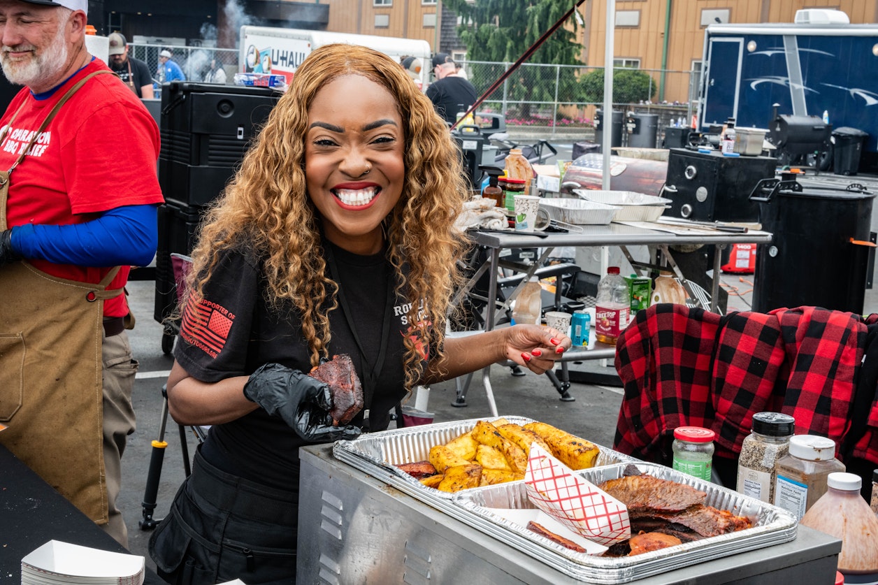A smiling vendor serves barbecue at an outdoor cook-off, holding a piece of smoked meat over trays filled with grilled pineapple and sliced brisket. Cooking equipment, smokers, and other participants are visible behind her, creating a busy festival atmosphere.