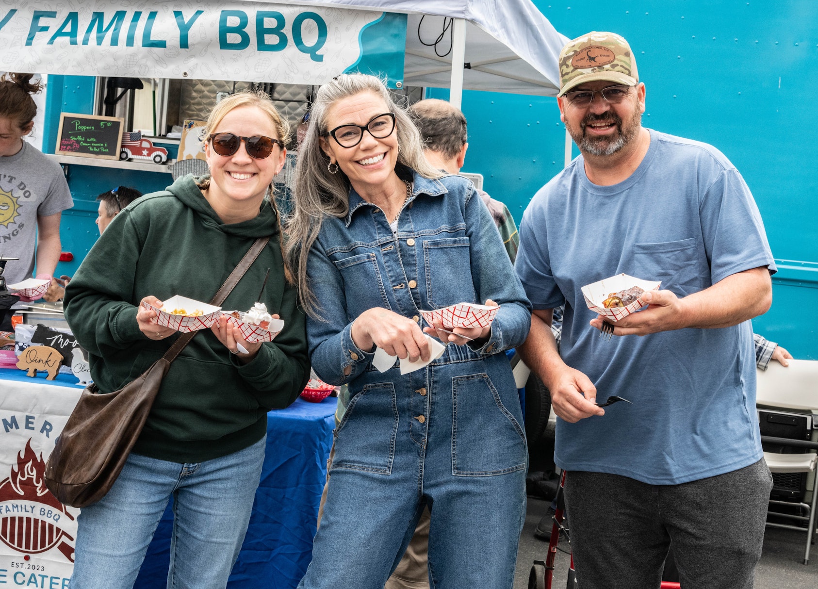 Three adults smile while holding small trays of barbecue food at an outdoor event. They stand in front of a food truck beneath a “Family BBQ” banner, with one person wearing a green hoodie, another in a denim jumpsuit, and the third in a blue T-shirt and baseball cap. The busy festival setting and vendor booth are visible in the background.