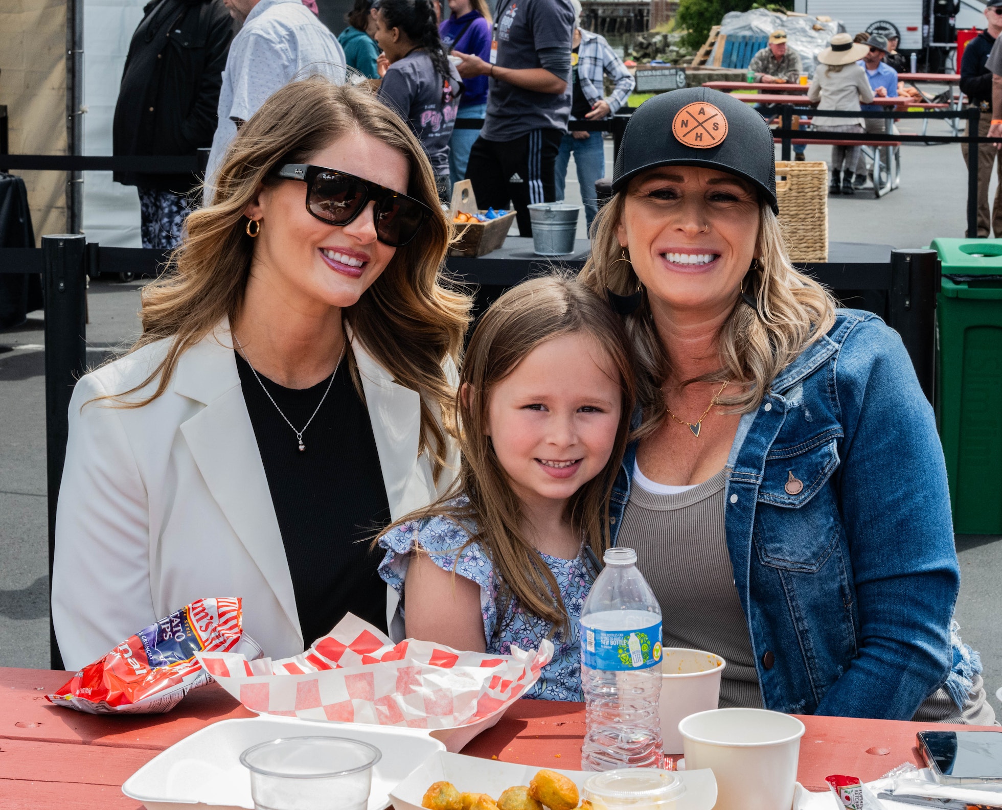 Three people sit together at the BBQ, Blues, Brews & Bourbon on the Bay, smiling at a picnic table with food and drinks. Two women, one wearing sunglasses and a light blazer and the other wearing a black hat and denim jacket, sit on either side of a young girl in a floral top. The crowd, tables, and vendor booths in the background suggest a lively festival atmosphere.