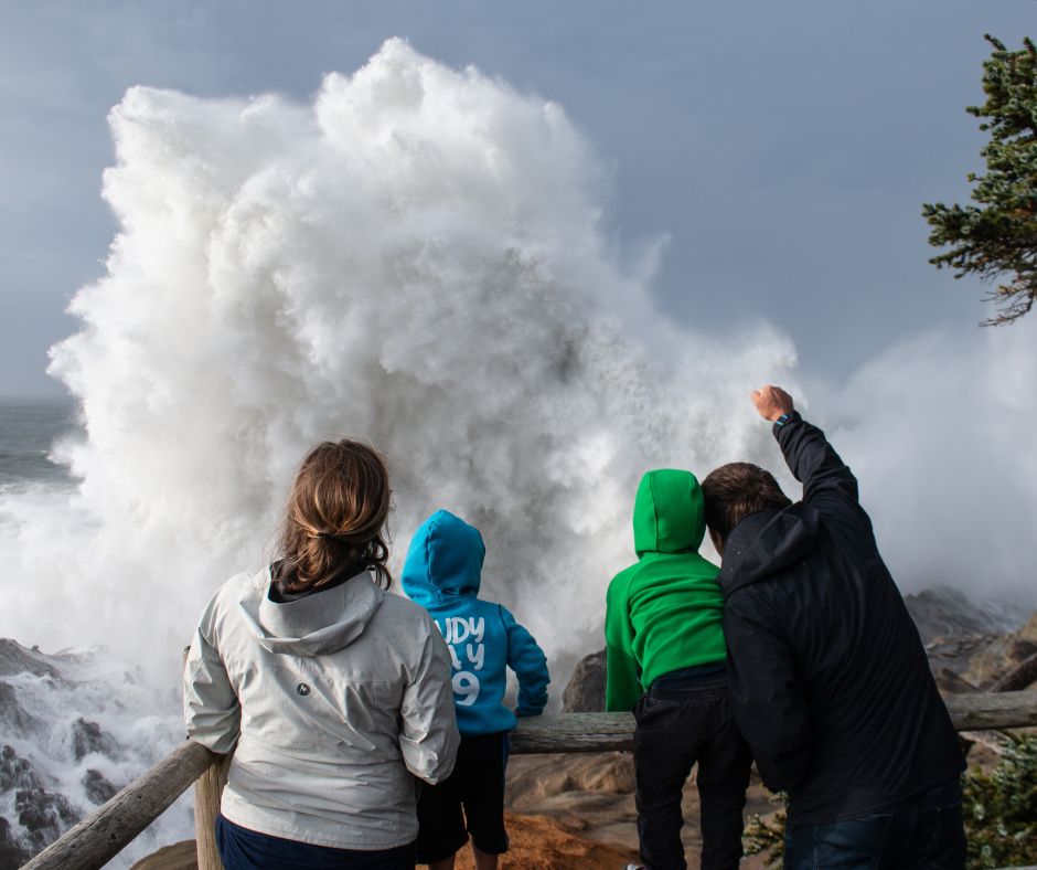A couple stands with their two young children at a designated lookout point in Shore Acres State Park, watching a massive winter-storm wave crash against the cliffs below. The family keeps a safe distance behind the viewing rail as the Pacific Ocean surges, sending spray high into the air against the rugged coastline.
