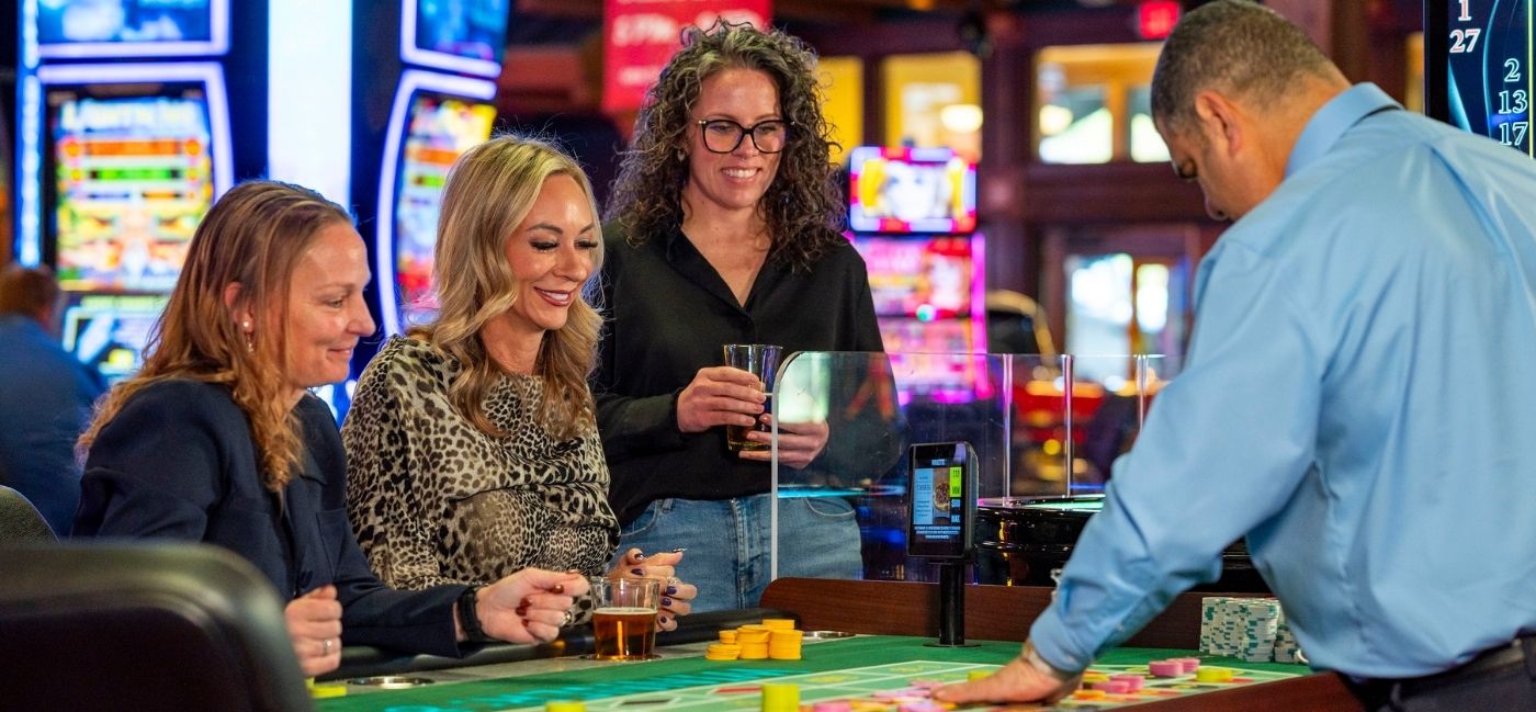 Three smiling women with drinks play roulette at a casino table while a dealer in a light blue shirt places chips on the felt. Brightly lit slot machines fill the background.