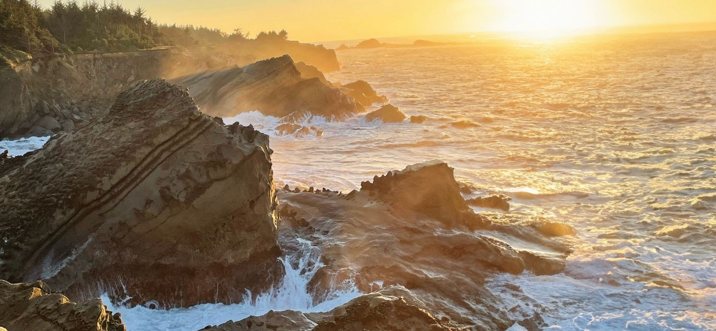 Golden sunset light washes over jagged, layered sandstone rock formations as waves crash and foam around them, with forested cliffs along the shoreline and the sun glowing low over the ocean.