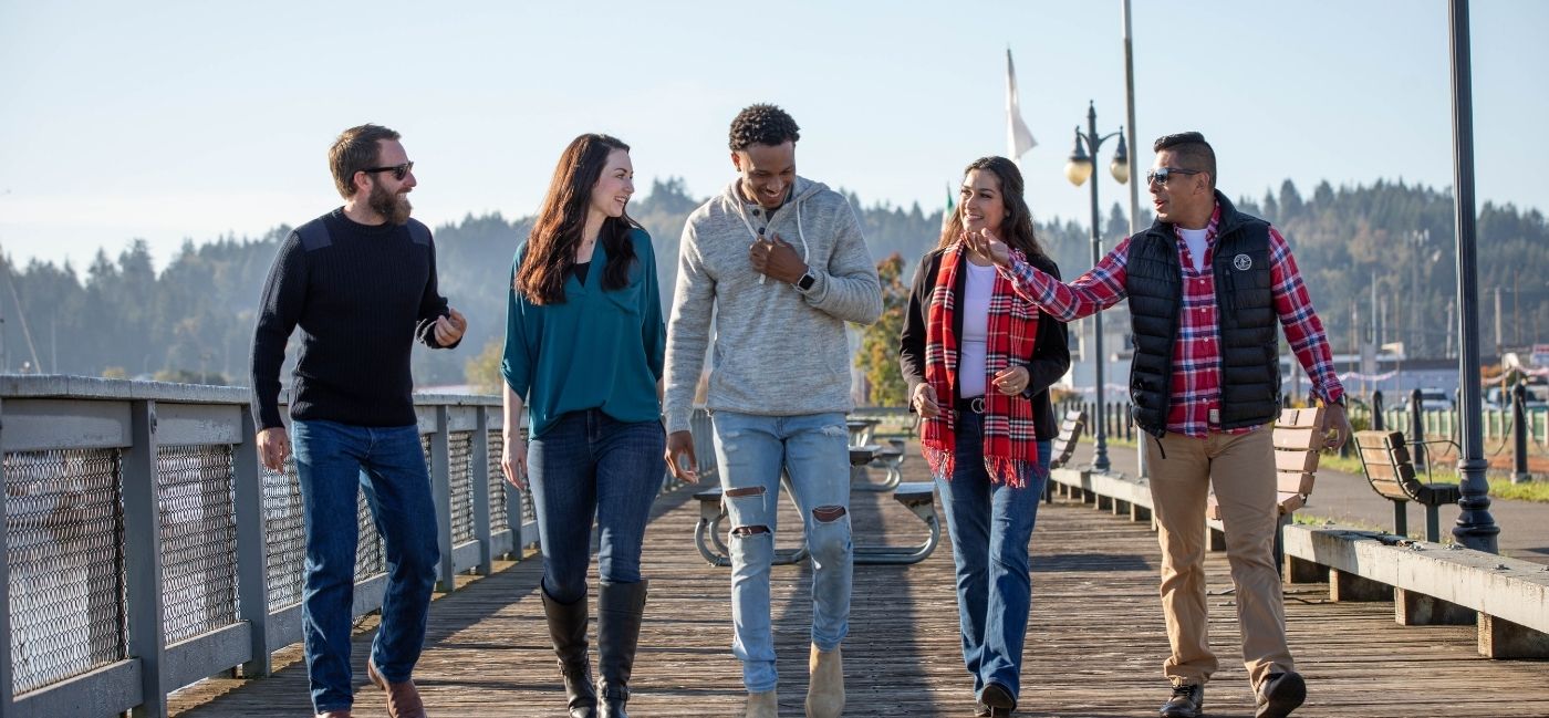 Five friends laugh and talk as they walk side by side along a sunny waterfront boardwalk, with benches, lampposts and forested hills in the background.