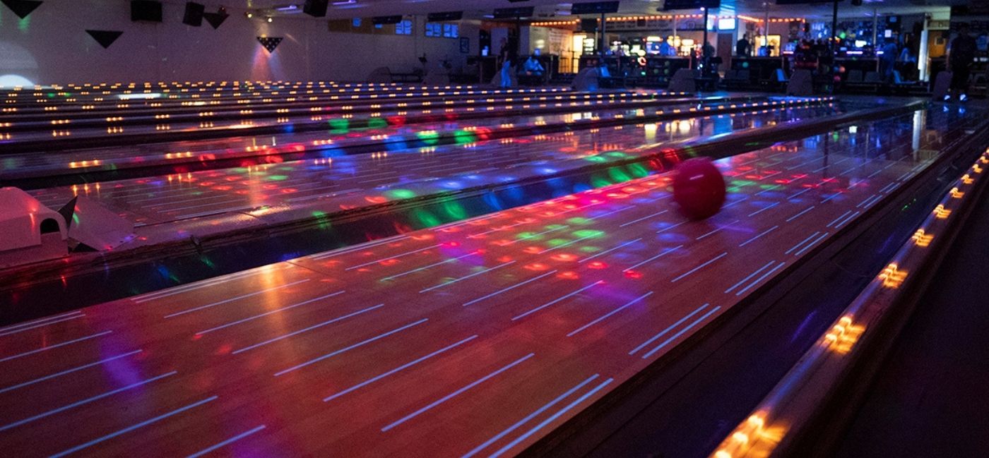 A red bowling ball rolls down a glossy lane during cosmic bowling, with colorful red, green and blue lights scattered across the lanes and glowing arrow markers and gutter lights in the dim room.