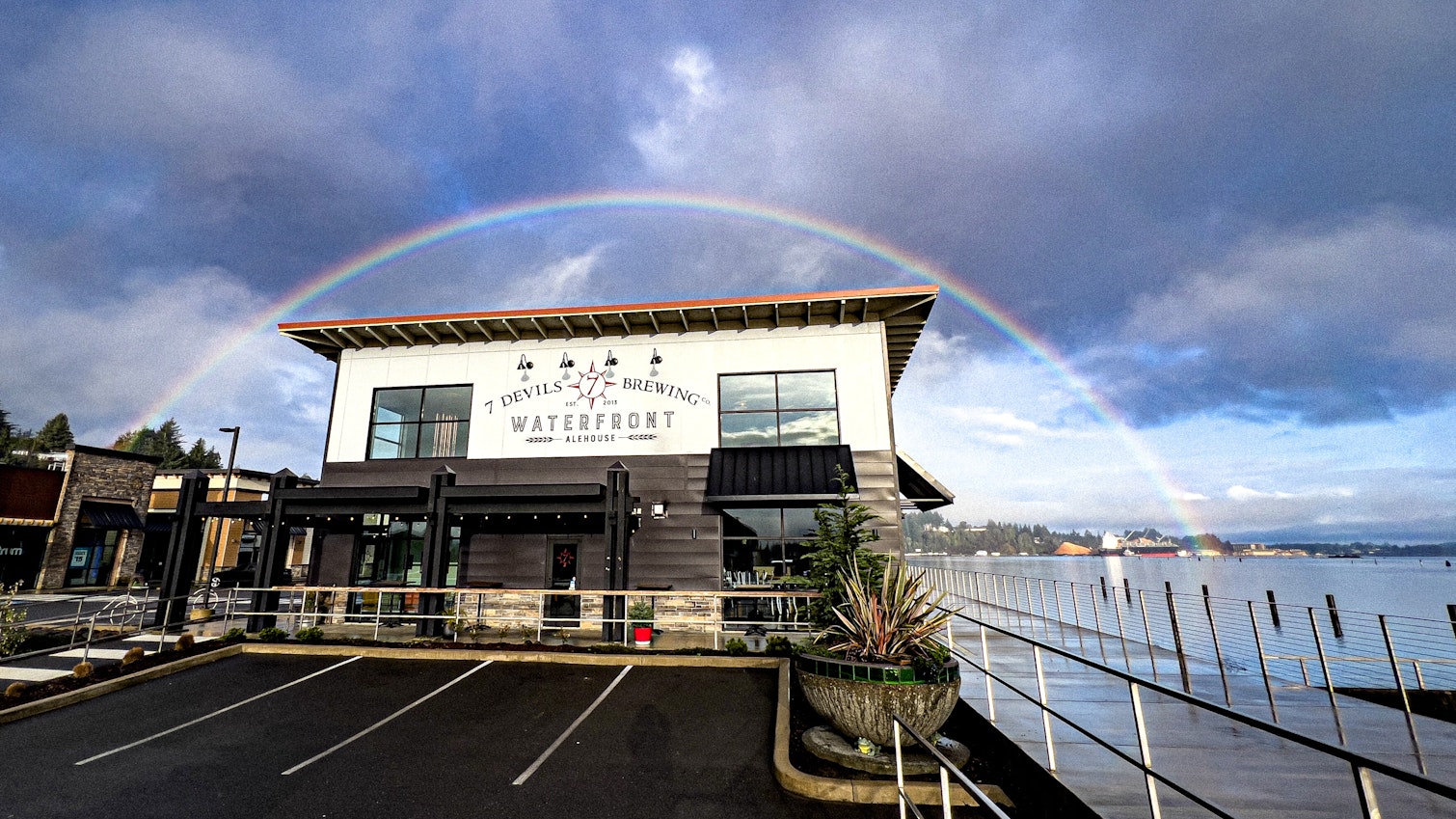 7 Devils Brewing Waterfront Alehouse along the bay with a rainbow arching overhead, cloudy skies, and a waterside walkway beside the building.