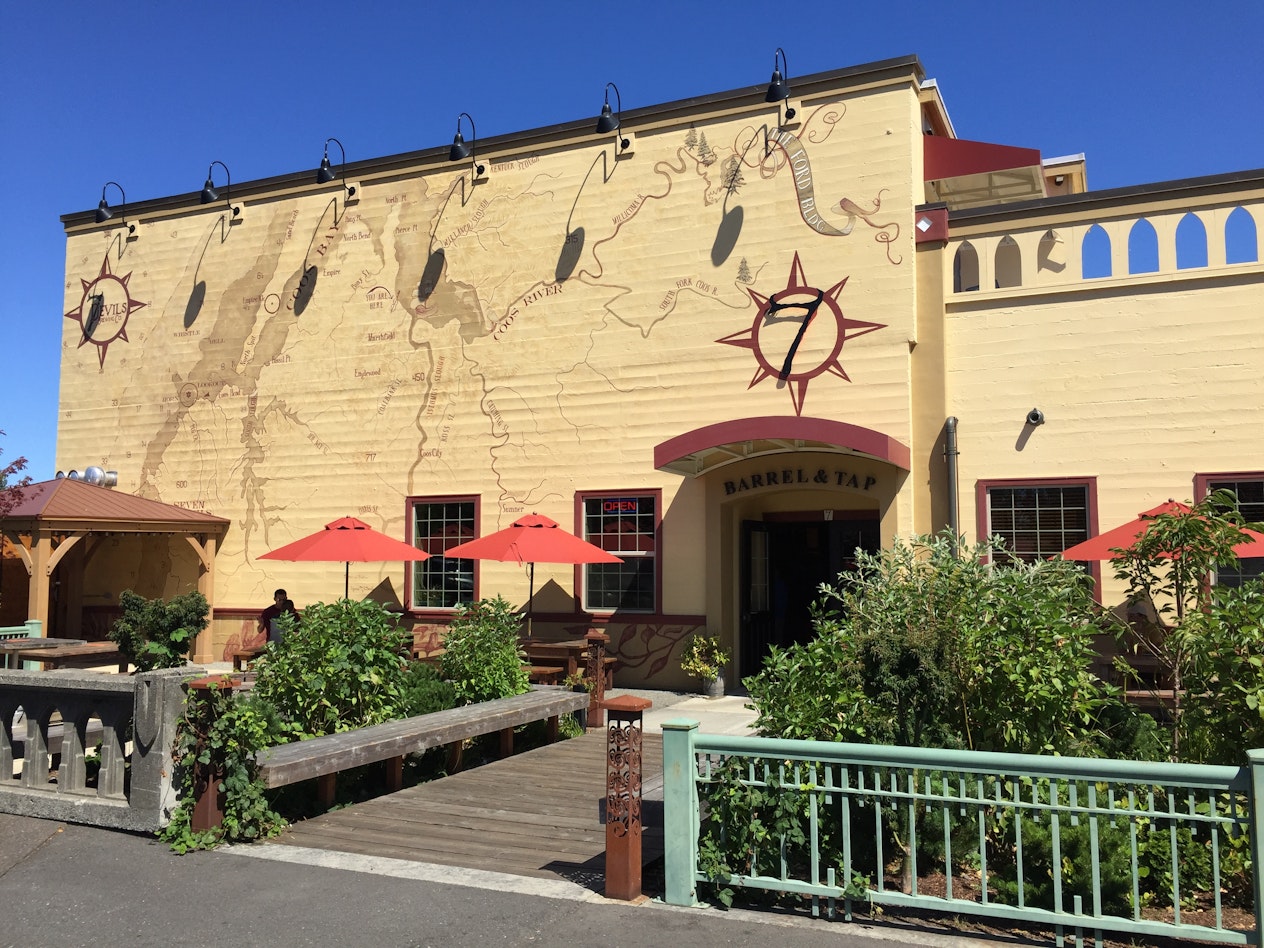 Exterior of 7 Devils Brewery in Coos Bay featuring a large mural map, outdoor patio seating with red umbrellas, and sunny blue skies.