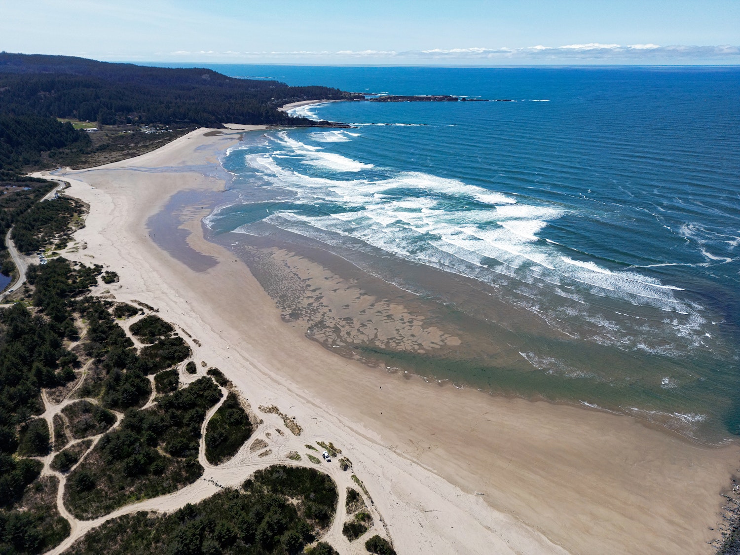 An aerial view of a wide, curved sandy beach along the Oregon Coast on a clear, sunny day. Waves with white surf roll in from the deep blue Pacific Ocean to the right, while sand dunes, scrubby vegetation, and a forested hillside line the left. A road and a few vehicles are visible near the dunes. A rocky headland and small coastal community are visible in the distance.