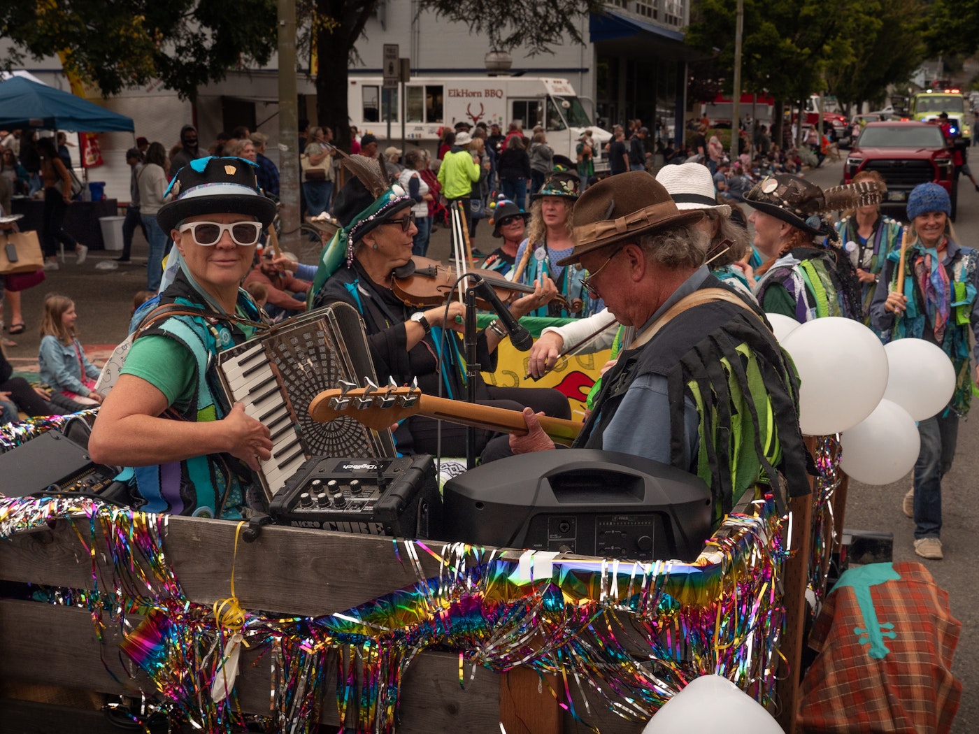 Colorfully costumed musicians playing accordion, violin, and guitar perform on a festive, tinsel-decorated float during the Bay Area Fun Festival parade in Coos Bay, Oregon, with crowds lining the street in the background.