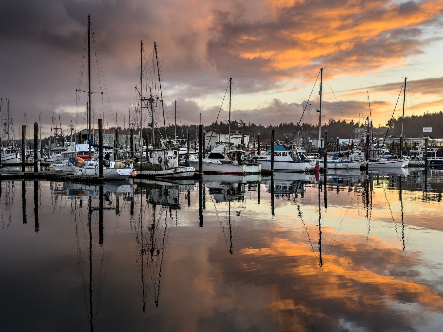 Fishing boats and sailboats moored at Charleston Harbor reflect on calm water at sunset, with dock pilings, masts, and a dramatic sky of layered clouds glowing above the working marina.