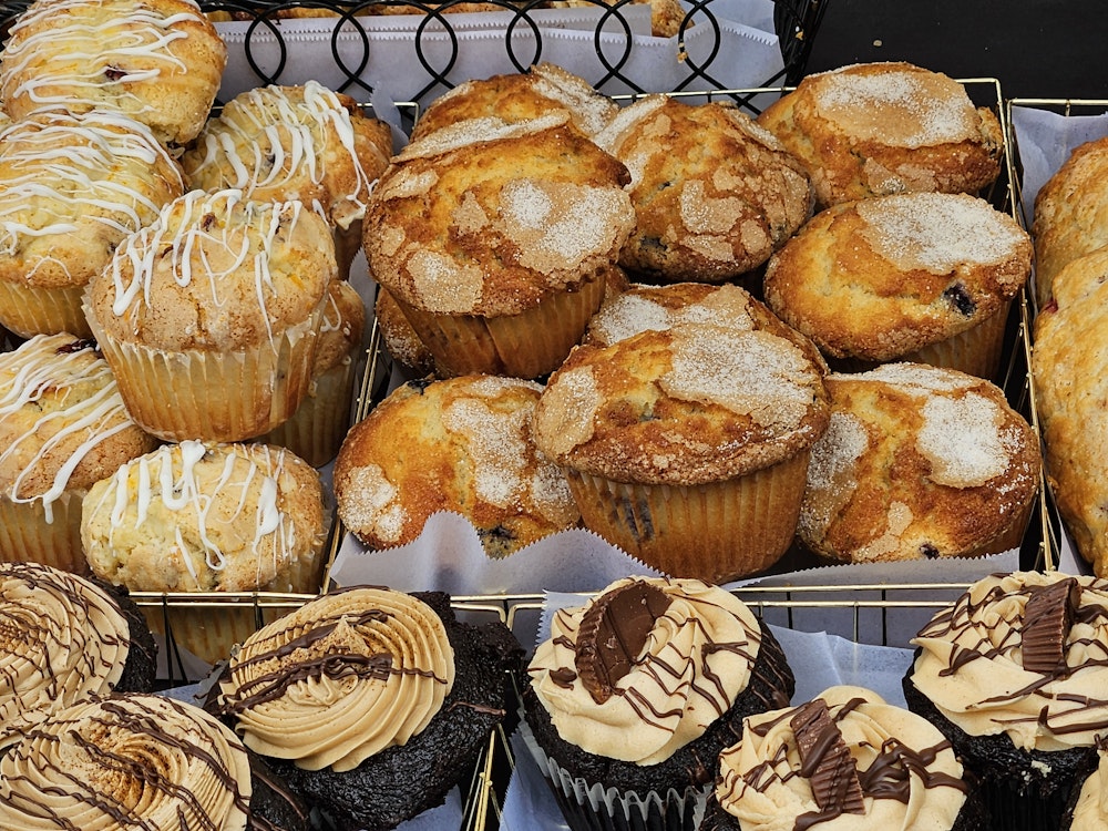 A bakery display filled with freshly baked muffins topped with sugar and icing, alongside chocolate cupcakes with swirled frosting and chocolate drizzle arranged neatly in trays.