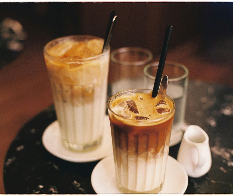 Two glasses of iced coffee with layered milk and espresso sit on white saucers, each with ice cubes and a black straw, with empty water glasses and a small creamer in the background on a café table.