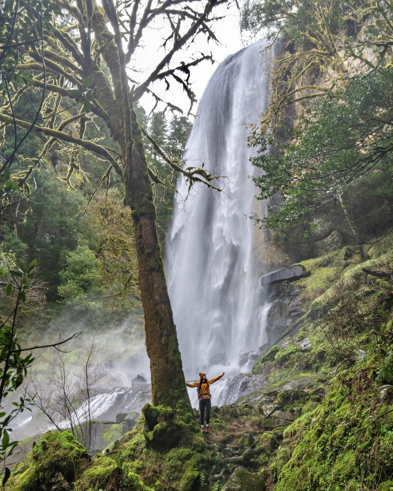 Ruby stands with her arms raised near the base of a waterfall in the Golden and Silver Falls Natural Area, surrounded by moss-covered rocks, mist, and lush forest vegetation.