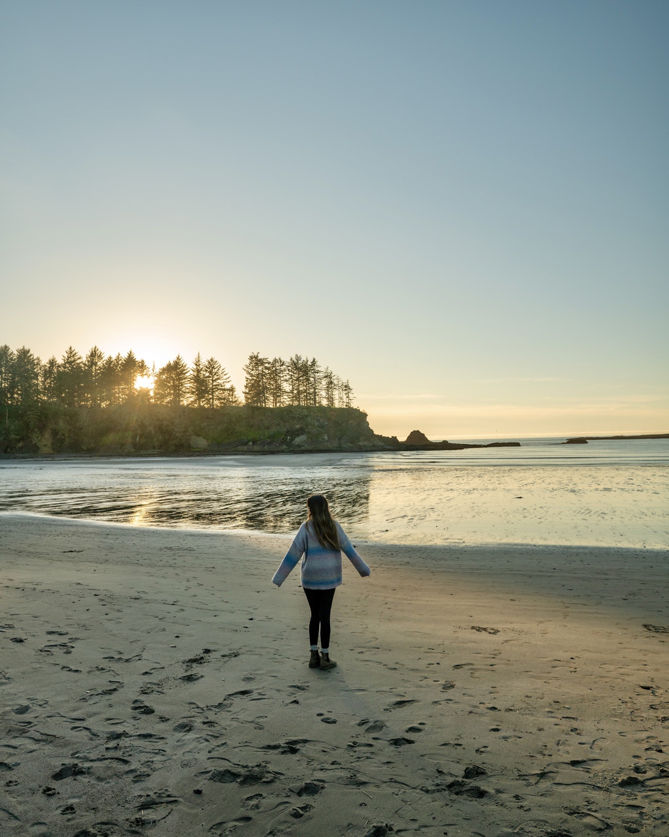 Ruby Tawes walks along a quiet beach at sundown during a visit to Sunset Bay State Park and stares at the calm water with trees in the background..