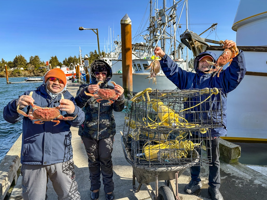 Three people standing on a dock at Charleston Marina hold up freshly caught Dungeness crabs beside a crab pot filled with traps and rope, with fishing boats and a working marina visible in the background on a clear day.