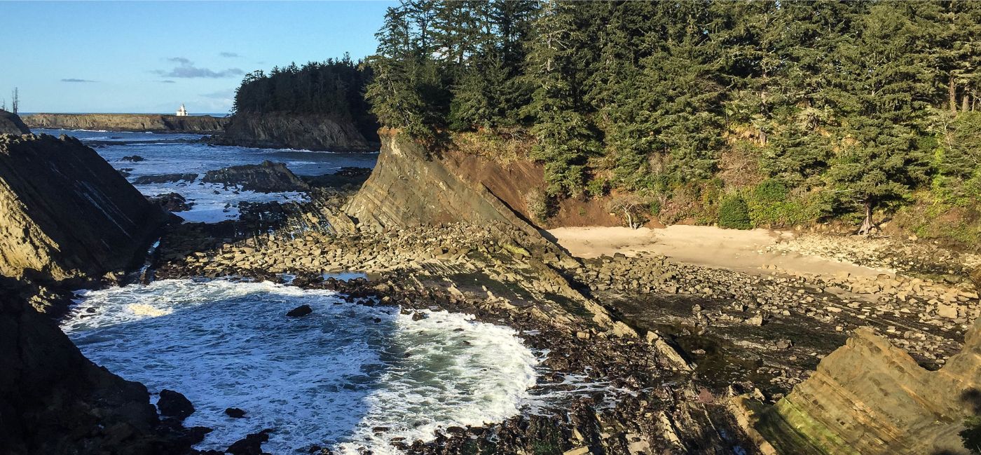 A rugged rocky cove at Shore Acres State Park on Oregon’s Adventure Coast, with waves crashing against layered rock formations, a small sandy beach nestled between the cliffs, dense evergreen forest above, and a lighthouse visible in the distance under a clear blue sky.
