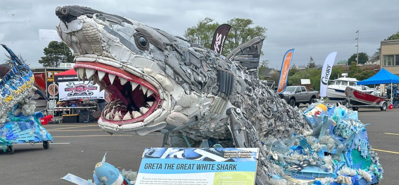 A large sculpture of a great white shark named Greta the Great White Shark, constructed entirely from recycled ocean plastic and debris, on display at an outdoor event on Oregon’s Adventure Coast, with an informational sign and vendor tents visible in the background.