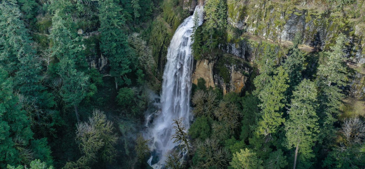 Aerial view of a tall waterfall cascading down a moss-covered rocky cliff face, surrounded by dense evergreen forest on Oregon’s Adventure Coast, with mist rising from the base of the falls.