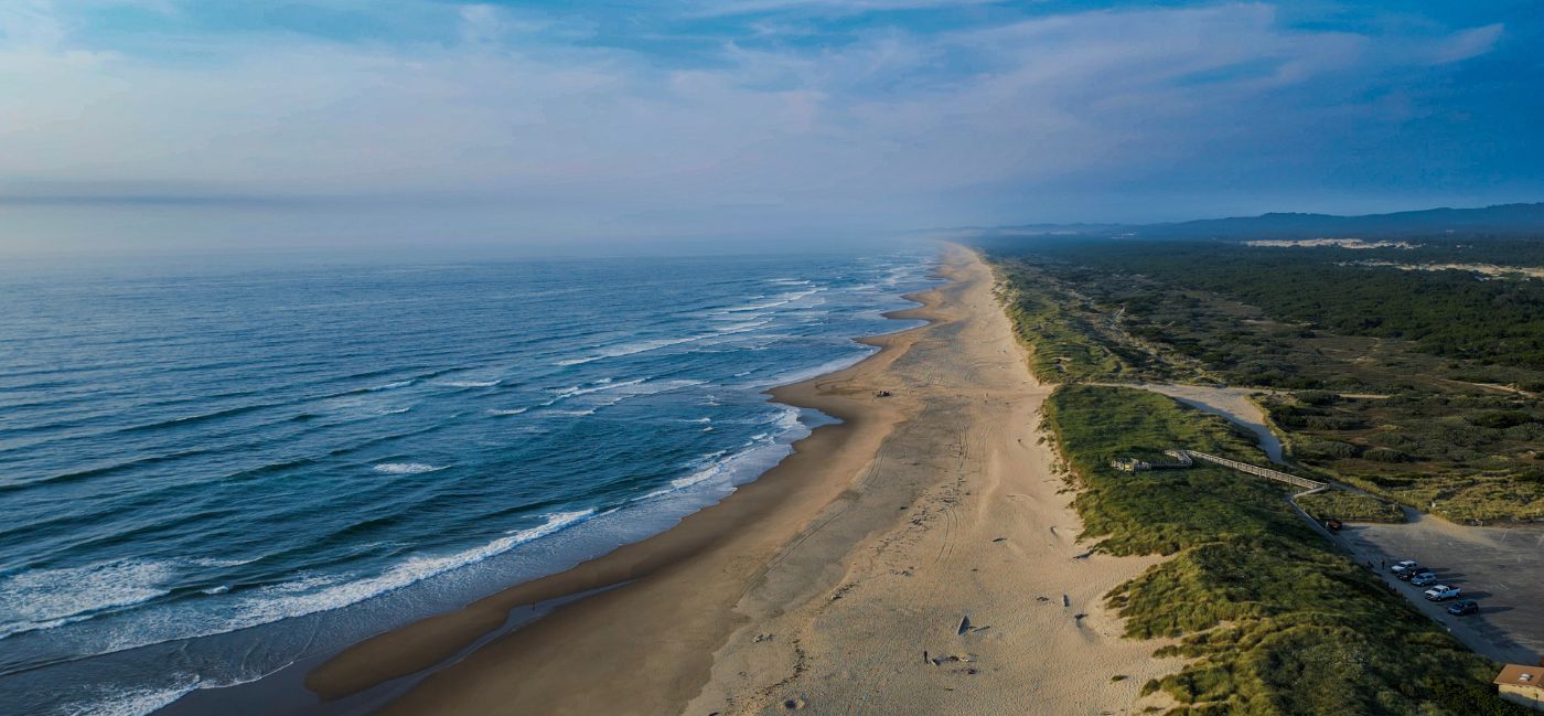 Aerial view of a long, wide sandy beach stretching into the distance along Oregon’s Adventure Coast, with rolling ocean waves on the left and green coastal dunes, forest, and a parking area on the right under a partly cloudy sky.