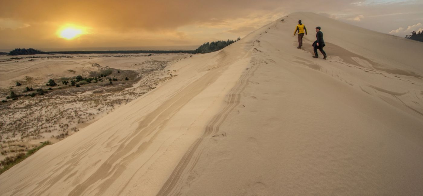 Two people hiking along the ridge of a large sand dune at sunset on Oregon’s Adventure Coast, with sweeping views of the coastline and forested landscape below under a golden orange sky.