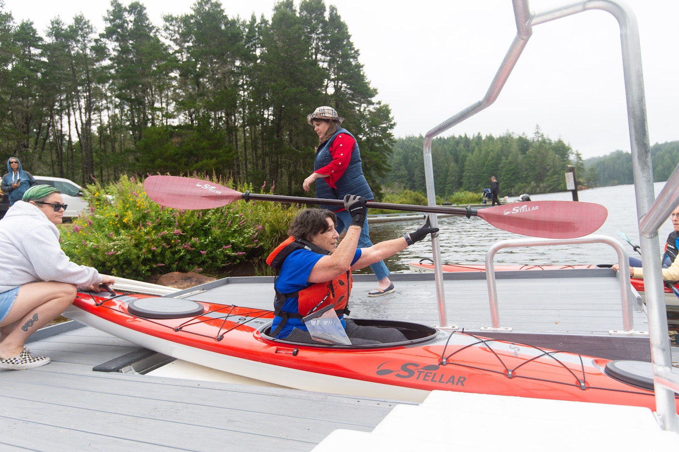 Person wearing a life jacket sits in a red kayak at a dock, holding a paddle while others assist with launching into a calm lake surrounded by trees.