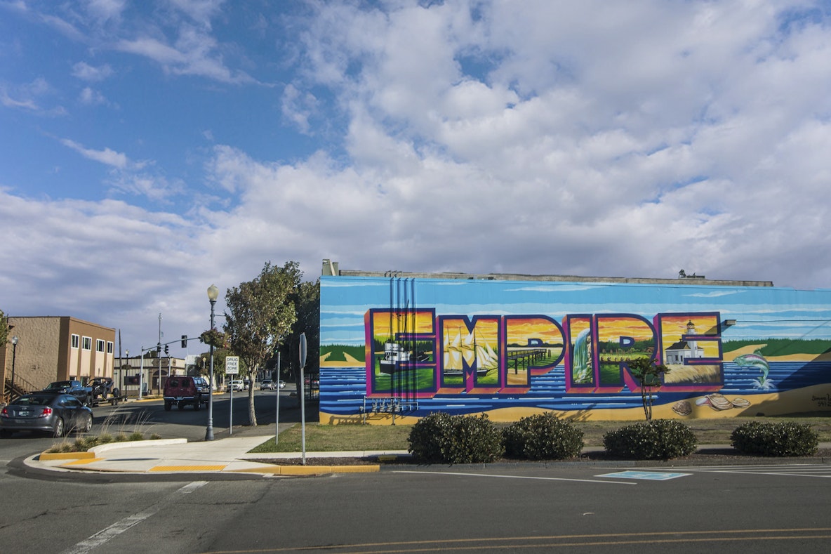 Large colorful “Empire” welcome mural painted on a building wall in the Empire District of Coos Bay, with each letter illustrating scenes of local history, waterfront views, and community landmarks along a street corner. Large colorful “Empire” welcome mural painted on a building wall in the Empire District of Coos Bay, with each letter illustrating scenes of local history, waterfront views, and community landmarks along a street corner.