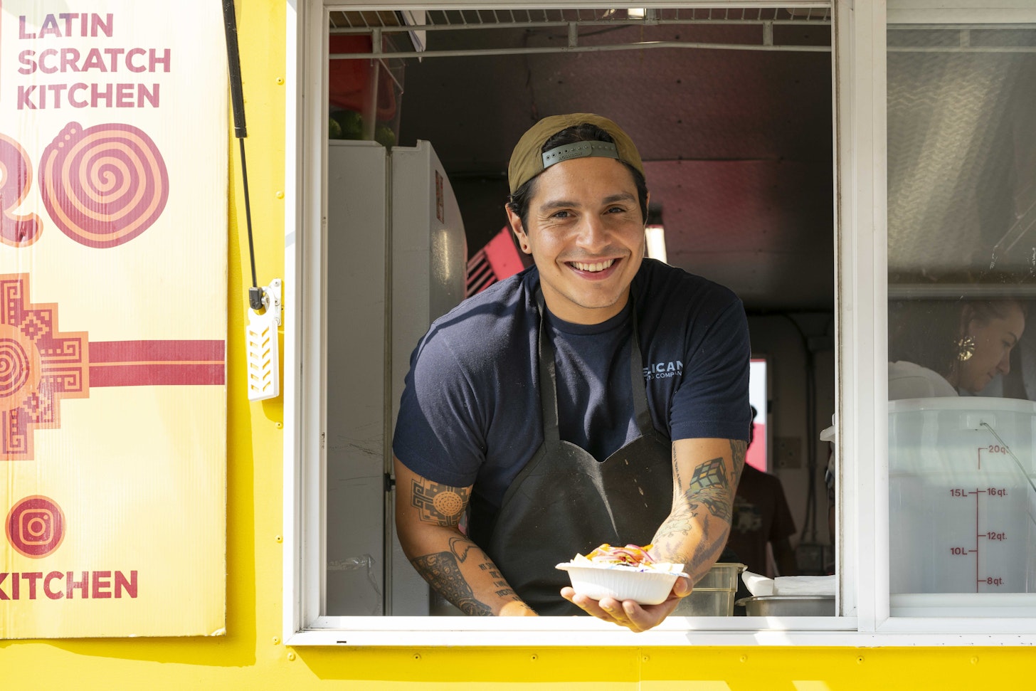 Image courtesy of Ko-Kwel Casino Resort | Coos Bay Smiling man with tattooed arms leans out the serving window of a yellow Latin Scratch Kitchen food truck, holding out a to-go container of food.