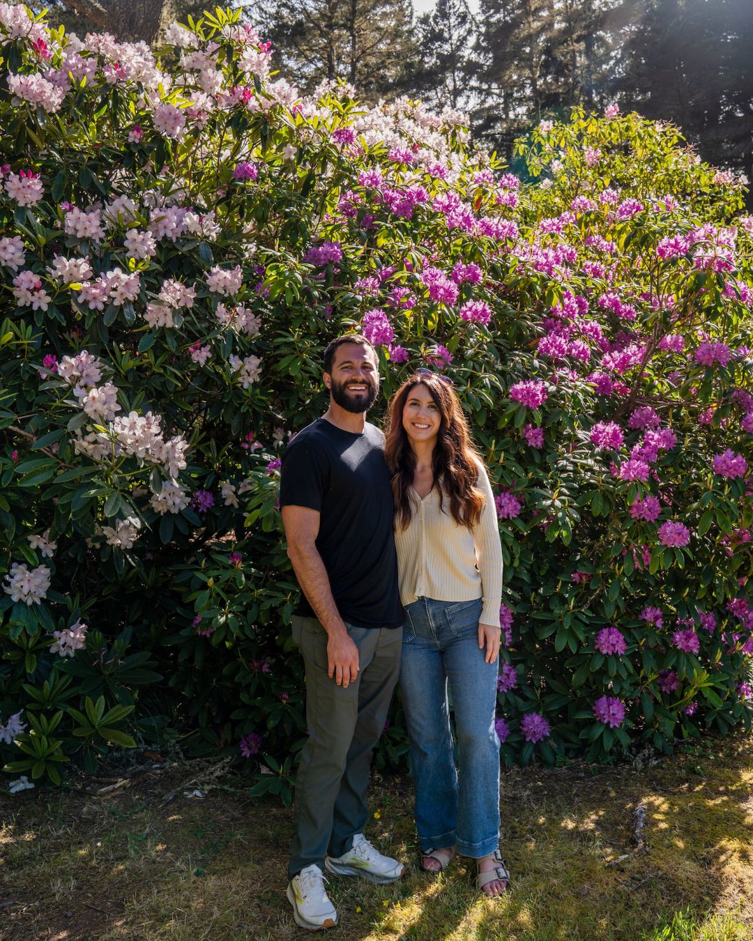 Photography by Adventuring Eyes Couple smiling in front of a large blooming rhododendron bush with pink and white flowers.