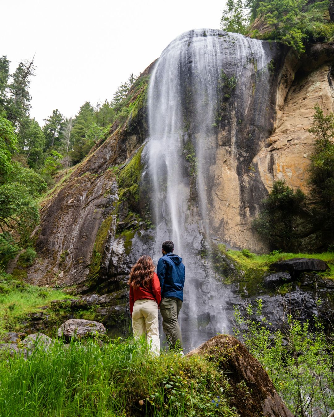 Photography by Adventuring Eyes Couple standing at the base of a tall waterfall cascading down a moss-covered rock cliff surrounded by forest.