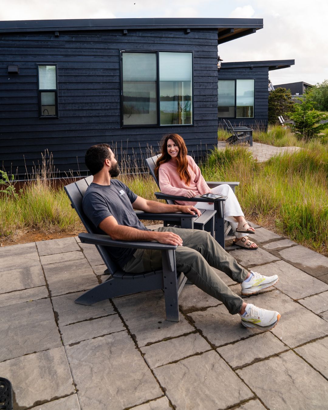 Photography by Adventuring Eyes Couple relaxing in Adirondack chairs on a stone patio outside a modern dark-sided cabin at Bay Point Landing.