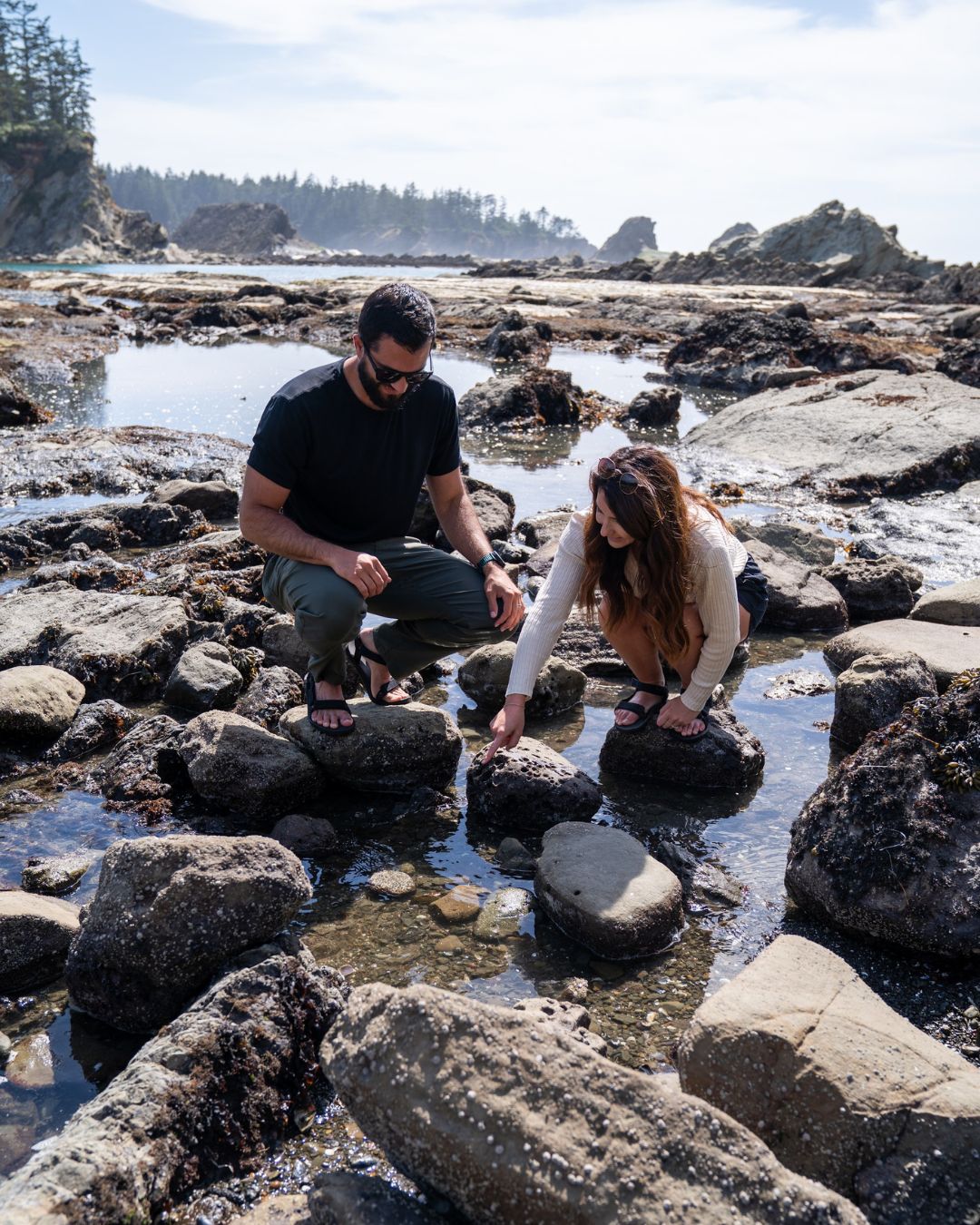 Photography by Adventuring Eyes Couple crouched among rocky tide pools on the coast, exploring the water with sea stacks in the background.