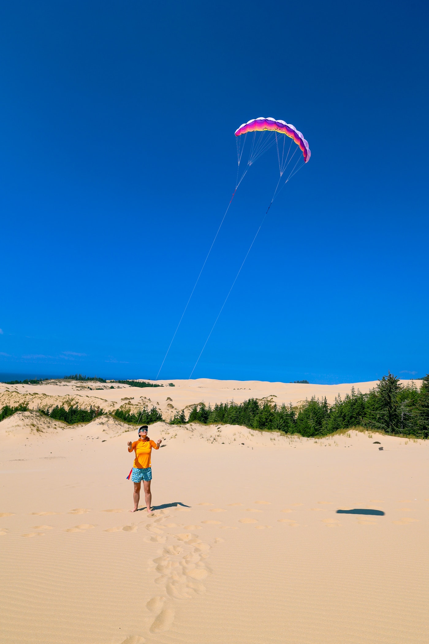 Person in orange shirt flying a colorful power kite on an open sand dune with forest and blue sky in the background.