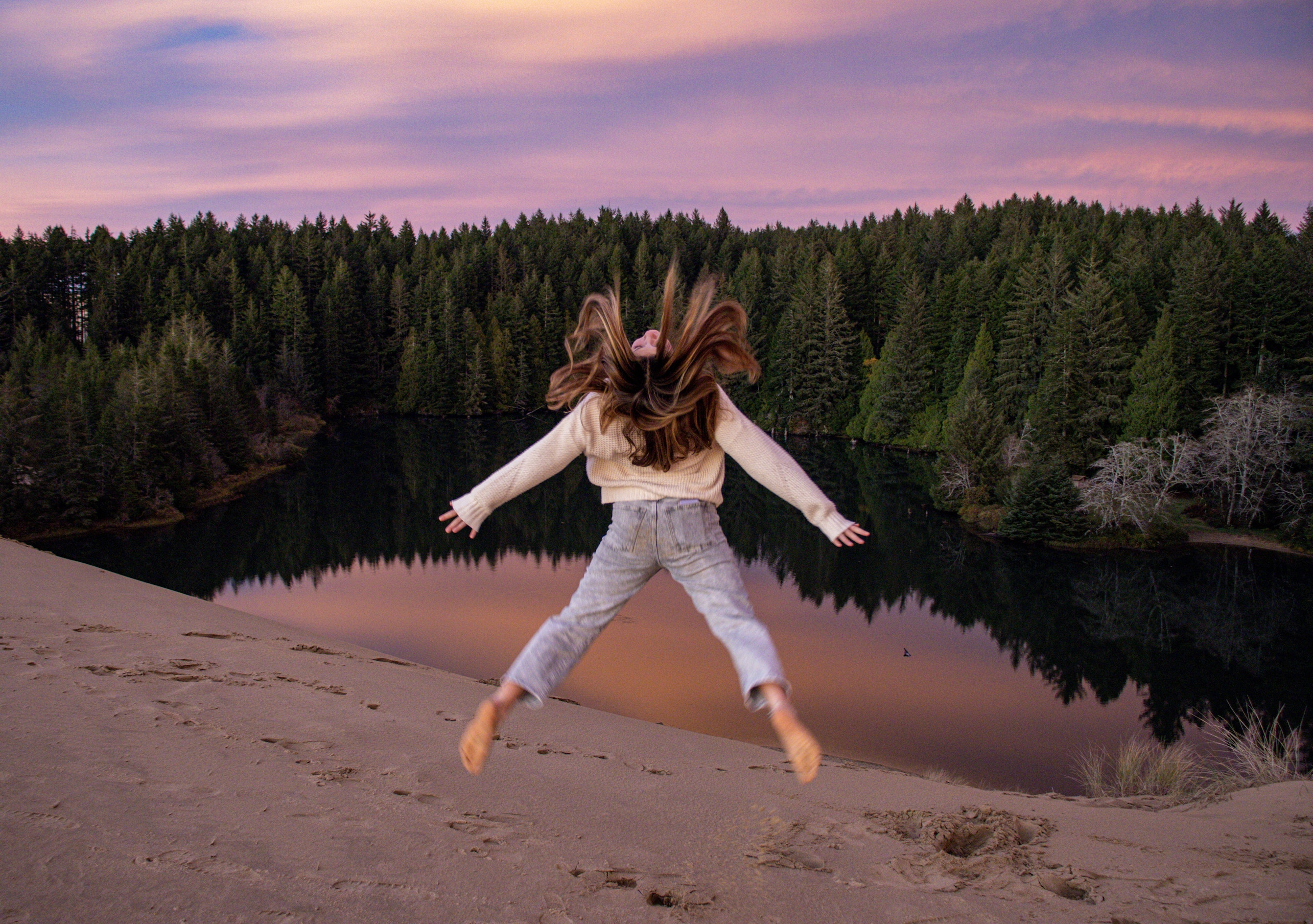 Woman jumping joyfully on a sand dune with hair flying, a still lake and pine forest reflected below at dusk.