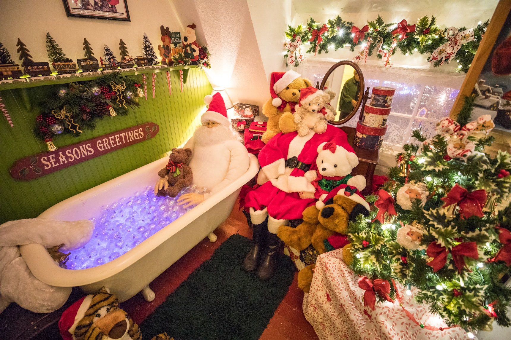 A whimsical holiday display inside The Garden House at Shore Acres features a Santa mannequin relaxing in a glowing bubble-filled bathtub beside another Santa figure surrounded by teddy bears, festive garlands, and a small decorated Christmas tree—one of the many creative scenes showcased during the Annual Holiday Lights at Shore Acres.