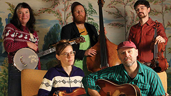 Five-piece string band posing for a promotional photo. Two members stand in back holding a banjo and upright bass; two stand to the sides holding a fiddle and banjo. One member sits in front holding an acoustic guitar. All wear casual patterned clothing in front of a painted nature backdrop.