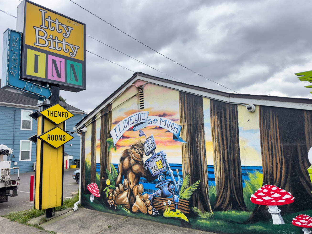 Exterior mural at the Itty Bitty Inn in North Bend, Oregon showing Bigfoot hugging a robot in a forest setting beneath a banner reading “I Love You So Much,” with the colorful Itty Bitty Inn sign visible nearby. Exterior mural at the Itty Bitty Inn in North Bend, Oregon showing Bigfoot hugging a robot in a forest setting beneath a banner reading “I Love You So Much,” with the colorful Itty Bitty Inn sign visible nearby.