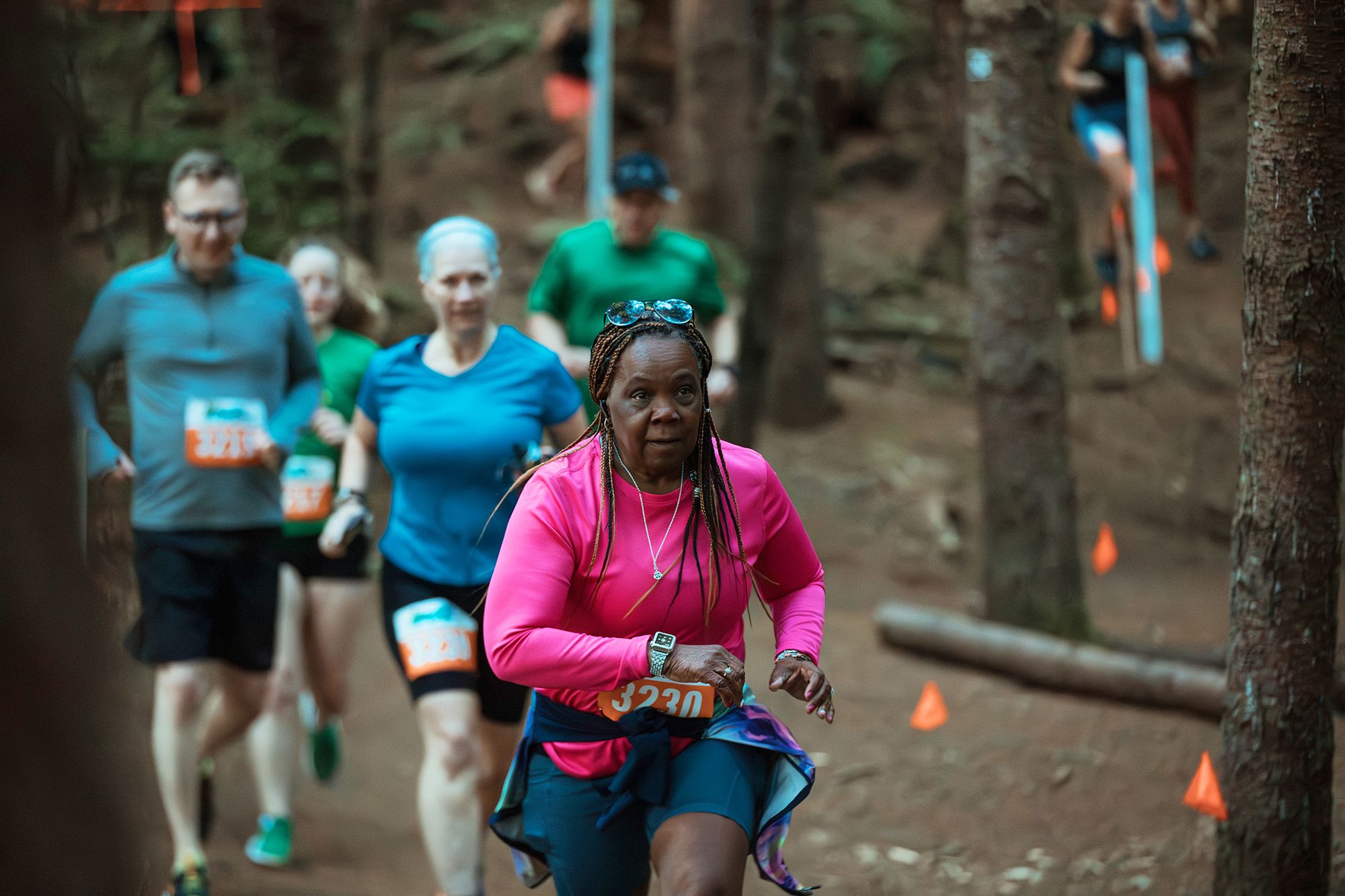 A group of runners moves along a wooded trail during a race, with a woman in a bright pink top leading the way as others follow behind among tall trees and marked course flags.