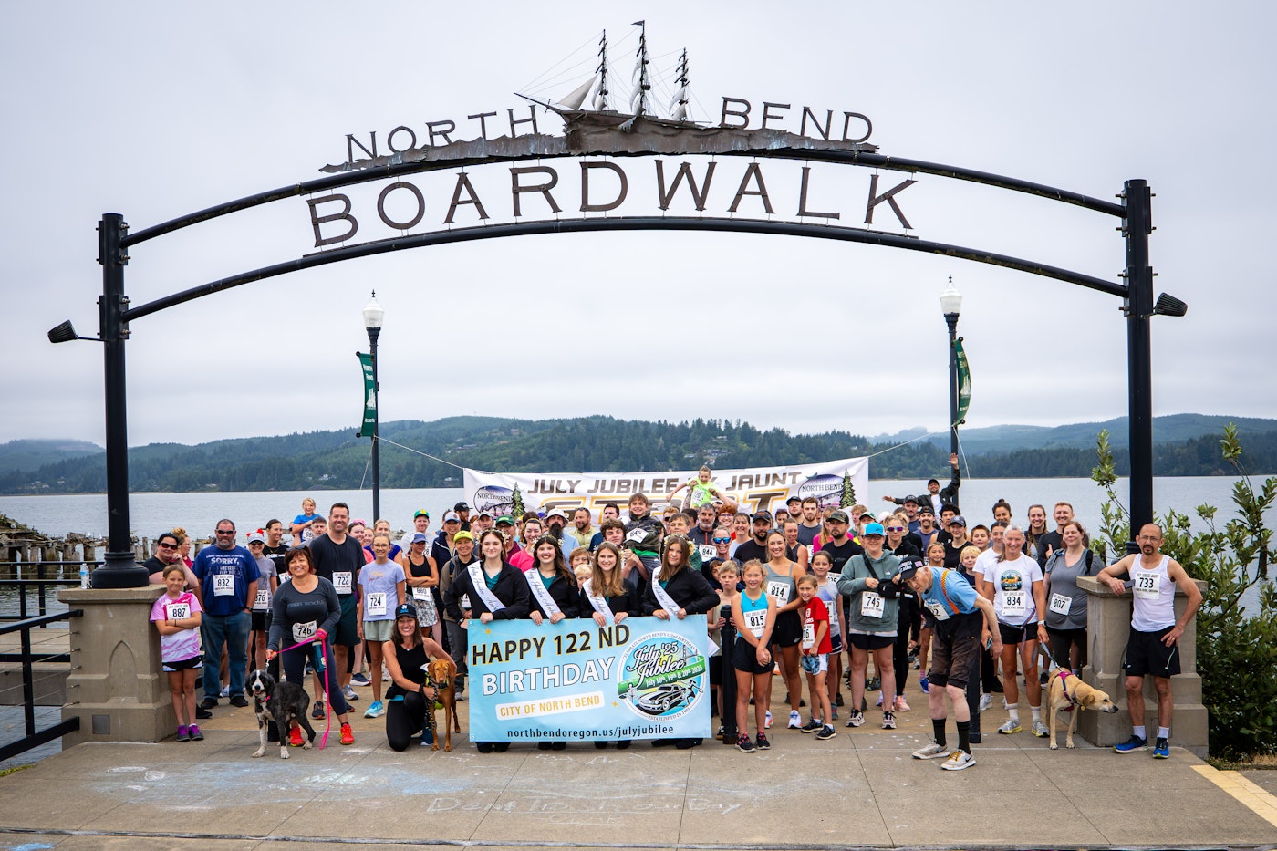 A large crowd of runners, walkers, and dogs posed for a group photo under the North Bend Boardwalk archway sign, with a bay and forested hills in the background, celebrating the city&rsquo;s 122nd birthday and the 2025 July Jubilee Jaunt 5K.