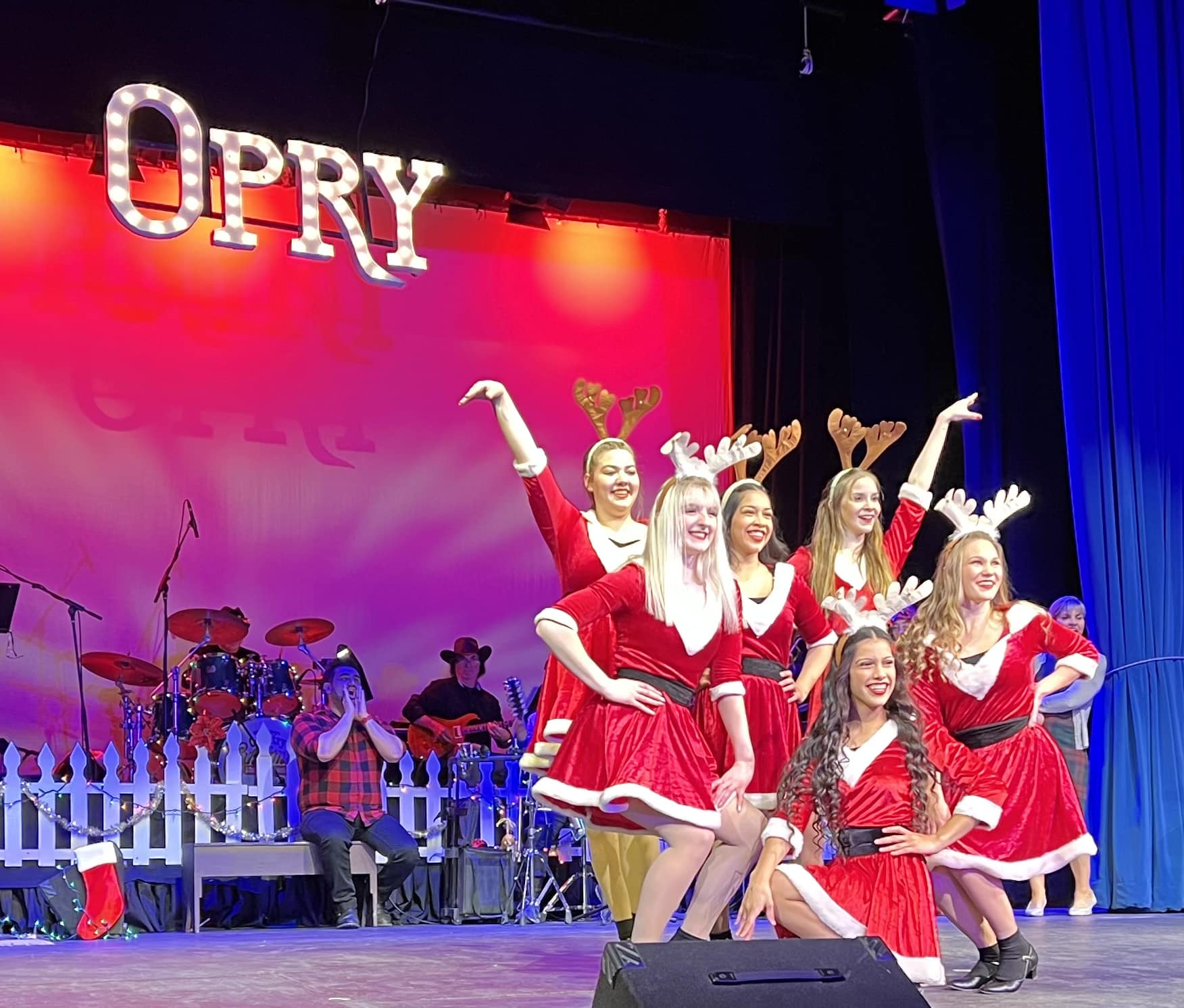 A group of performers in red holiday dresses and reindeer antlers pose onstage during the Christmas Opry on the Bay at the Liberty Theatre, with the band playing behind them under a lit “OPRY” sign.