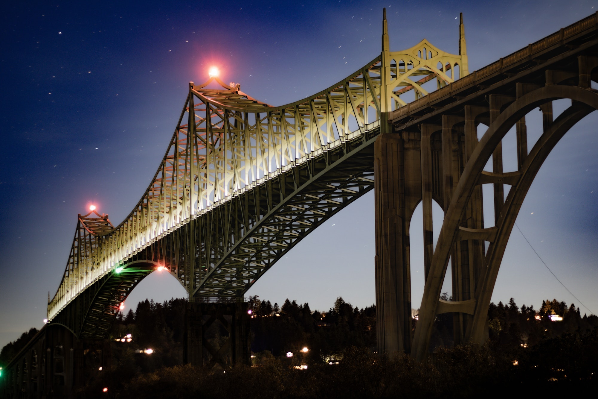 The  Conde B. McCullough Memorial Bridge in North Bend, a steel arch truss bridge, lit up at night against a starry twilight sky with silhouetted trees below.