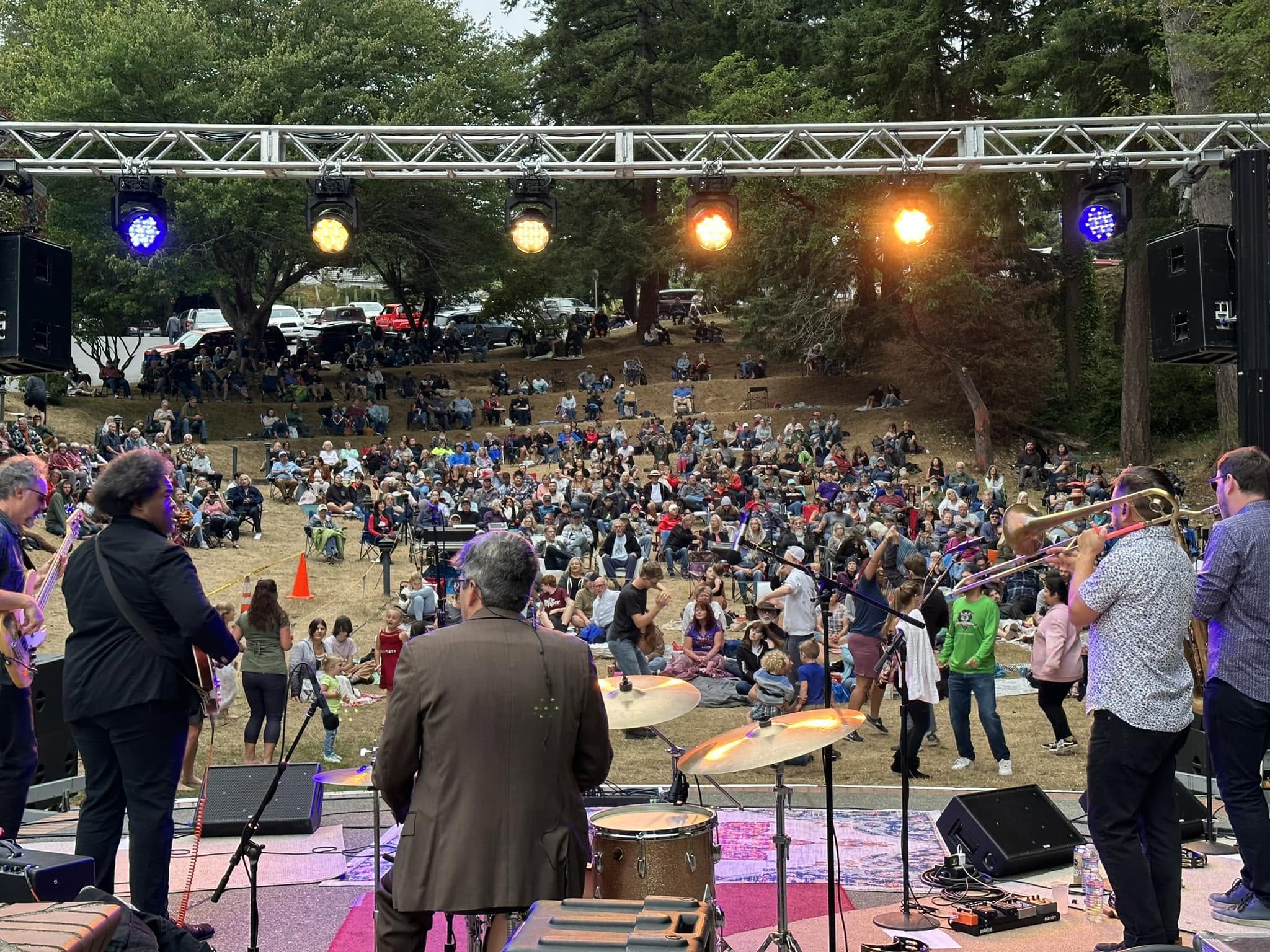 Photo courtesy of Music on The Bay View from the stage during one of the Music on the Bay performances in Mingus Park of a live band performing with guitars, trombone, and drums, facing a large crowd seated and dancing on a grassy outdoor hillside.