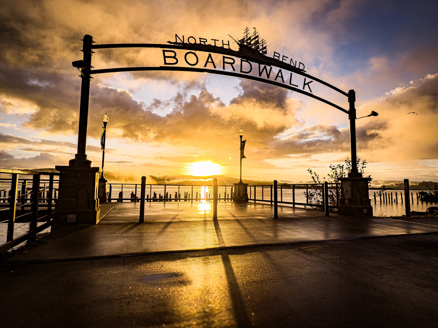 The North Bend Boardwalk archway silhouetted against a vivid golden sunrise over the water, with wet pavement reflecting the glowing sky.