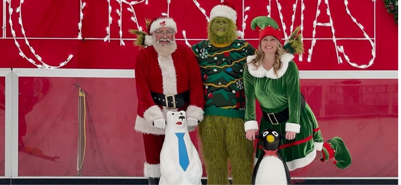 Santa, the Grinch and an elf pose on the ice with polar bear and penguin skating aids in front of a lighted Christmas sign.