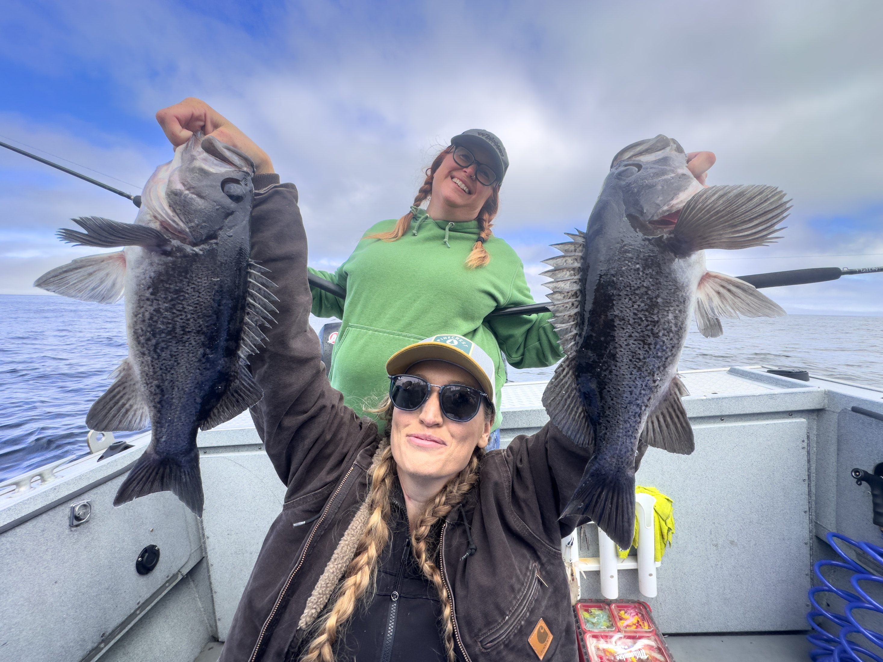 Two women on a fishing boat smiling and holding up rockfish they caught, with a cloudy sky and ocean in the background.