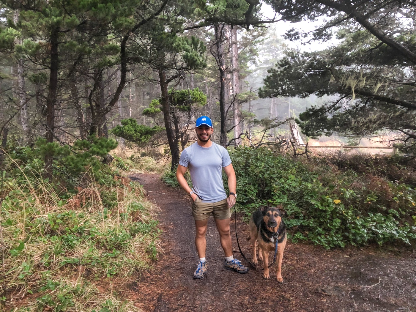Man in a blue cap standing with a leashed dog on a misty, pine-lined coastal trail.
