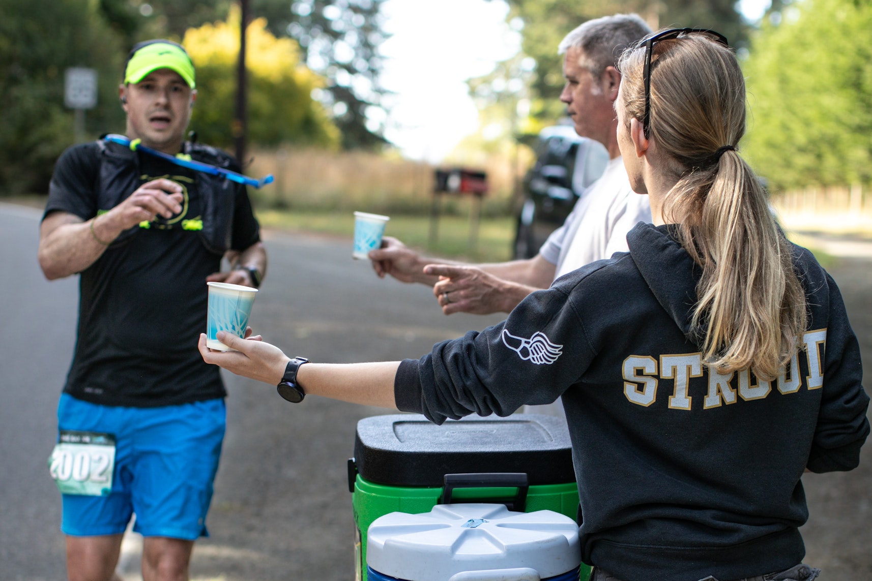 A runner wearing a hydration vest approaches a roadside aid station, reaching for a paper cup of water offered by volunteers standing beside a cooler during a race.