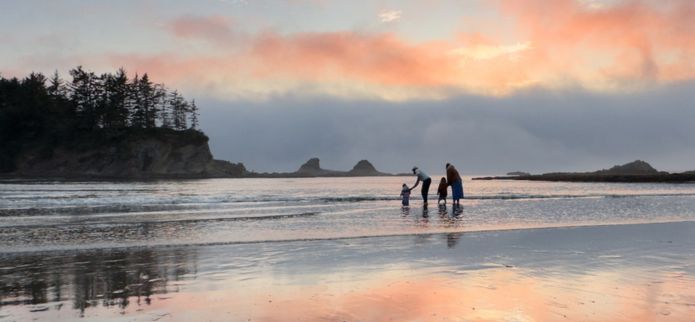 A family of three walks along a wide sandy beach at sunset, with soft pink and orange clouds reflecting on the wet shoreline and rocky sea stacks rising in the distance.