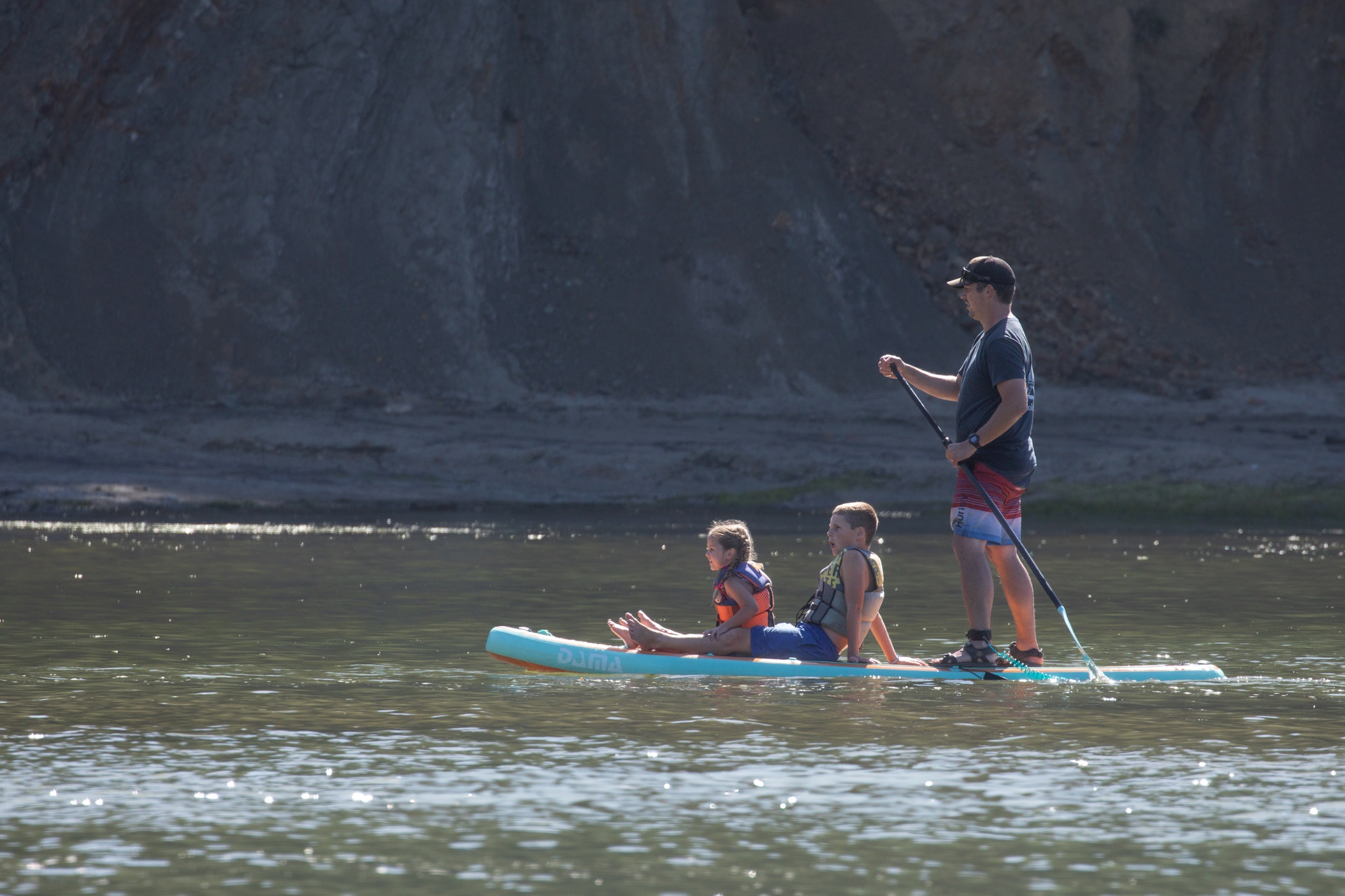 Man stands paddling a stand-up paddleboard in the calm waters below a steep rocky bluff at Sunset Bay State Park in Coos Bay while two children in life vests sit in front of him.