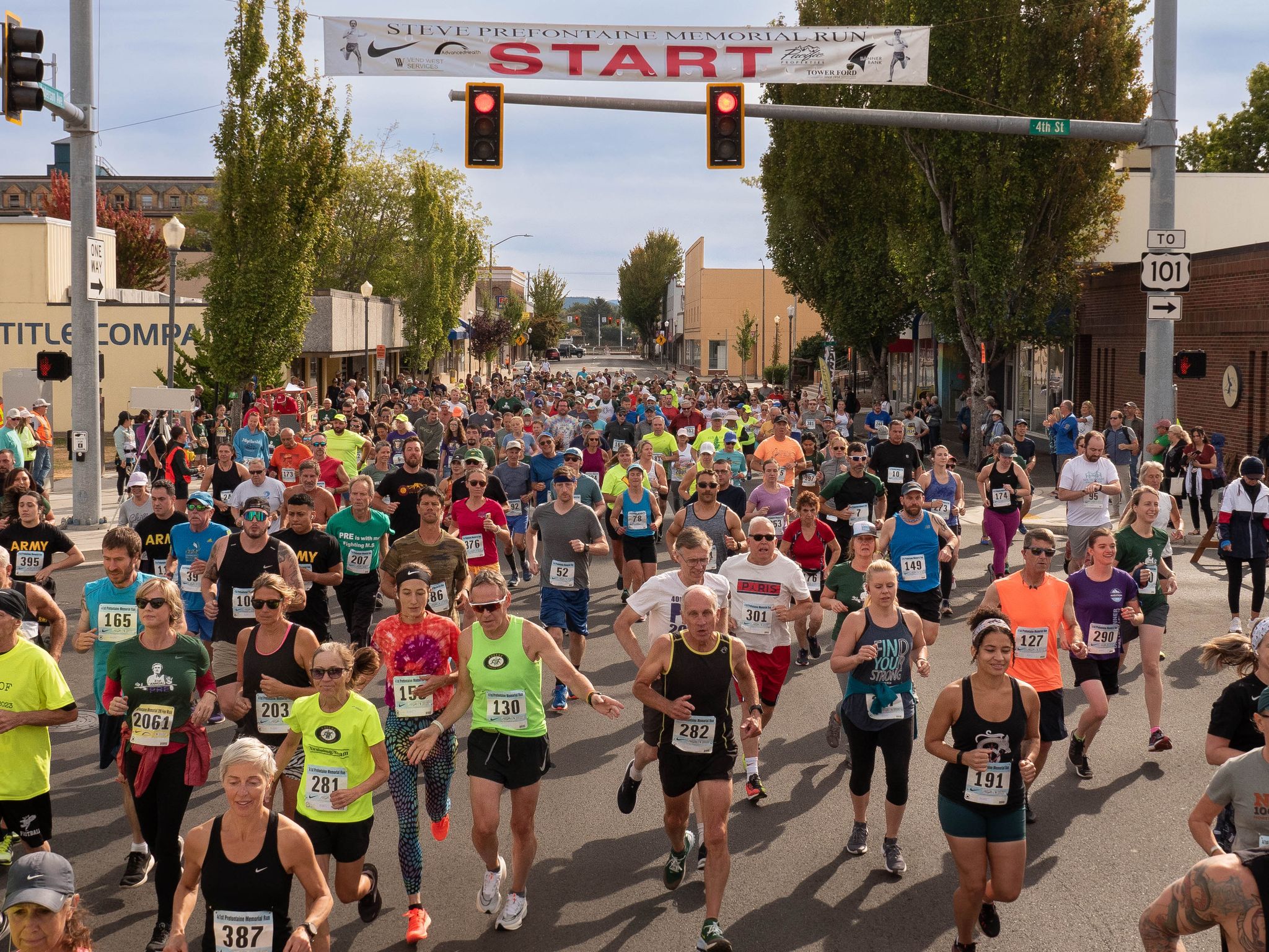 A large group of runners begins a race in a downtown street, passing under a “Steve Prefontaine Memorial Run” start banner as spectators line the sidewalks.